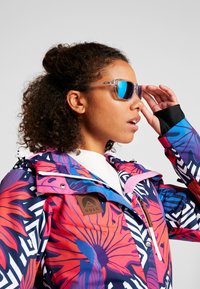 Bunte Skijacke mit floralen und geometrischen Mustern in Pink, Blau und Weiß, mit einem Logo-Patch und Reißverschlusstaschen. Klare Sonnenbrille inklusive.