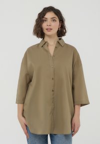 Camicia button-up khaki con vestibilità rilassata, maniche corte e colletto a punta. Texture liscia con bottoni marroni e spacchi laterali.