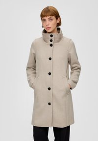Beige wollen jas met een hoge geribbelde kraag, sluiting met knopen aan de voorkant, zijzakken en zwarte knopen. Heeft een subtiele textuur en een getailleerde pasvorm.