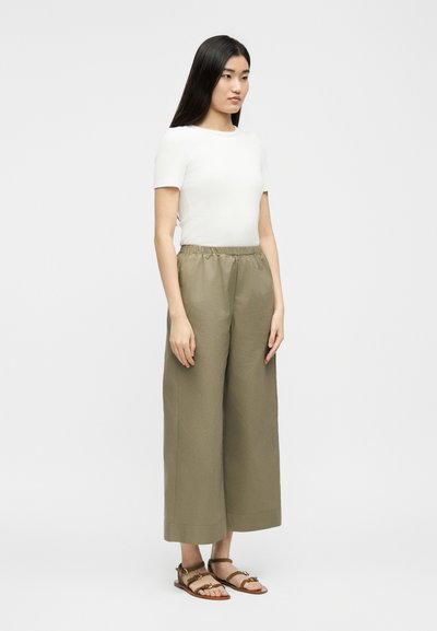 Νεαρή γυναίκα με μακριά σκούρα μαλλιά, που φοράει λευκό κοντομάνικο τοπ, φαρδιά, χακί παντελόνια και καφέ σανδάλια, στέκεται πλαγίως.