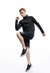 Schwarzes langärmliges Oberteil mit Reißverschlusskragen, kombiniert mit schwarzen Shorts. Das Outfit hat weiße Akzente und das Model trägt schwarze Sneaker.