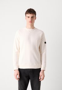 Lichtcrème sweatshirt met ribbeltextuur, ronde halslijn en lange mouwen. Bevat een klein zwart label op de linker mouw. Gecombineerd met donkere jeans.