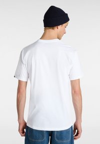Hvid kortærmet t-shirt vist fra ryggen, med en rund halsudskæring og en blød bomuldsstruktur, parret med en mørk strikket hue.