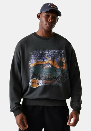 Uomo che indossa una felpa scura dei Los Angeles Lakers con stampa di un campo da basket e un cappellino abbinato, in piedi davanti a uno sfondo semplice.