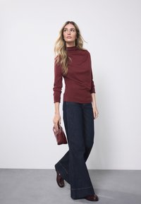 Maroon turtleneck top paired with dark blue flared jeans, holding a small maroon handbag. Smooth fabric textures and simple design elements.