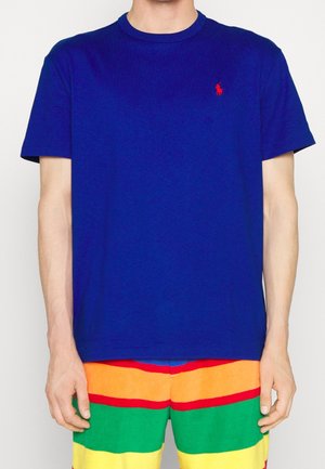Personne portant un T-shirt bleu uni avec un petit logo rouge sur la poitrine, assorti à un short rayé multicolore en orange, rouge, vert et jaune.