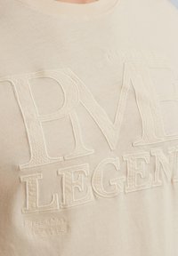 Beige puuvillane t-särk, mille esiküljel on tekstureeritud, brodeeritud ülemõõduline "PME LEGEND" logo, millel on õrnad õmblusdetailid ja ümmargune kaelus.