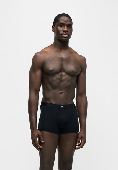 Homme musclé à la peau foncée, debout sur un fond uni, portant un boxer noir avec un petit logo sur la ceinture.