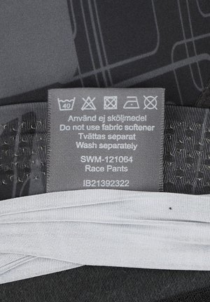 Løbebukser i sort stof med et struktureret mønster. Plejelabel viser vaskeanvisninger og produktdetaljer i grå.