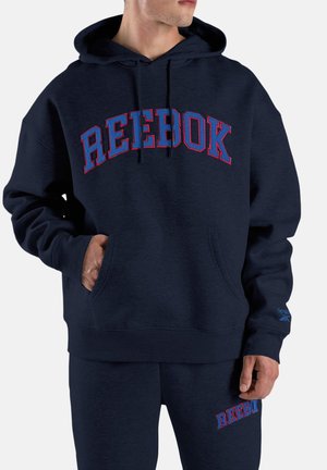 Hombre vistiendo sudadera con capucha azul marino de Reebok y pantalones deportivos a juego, con el logo de Reebok destacado en ambas prendas.