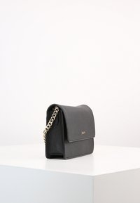Borsa a spalla in finta pelle nera con tracolla a catena, forma rettangolare, texture liscia e chiusura a pattina frontale. Dettaglio del logo dorato visibile.