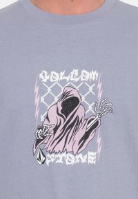 Lichtpaarse T-shirt met een grafisch ontwerp van een in een cape geklede figuur met schedelhandschoenen, omringd door bliksemschichten en de tekst "Volcom Stone."