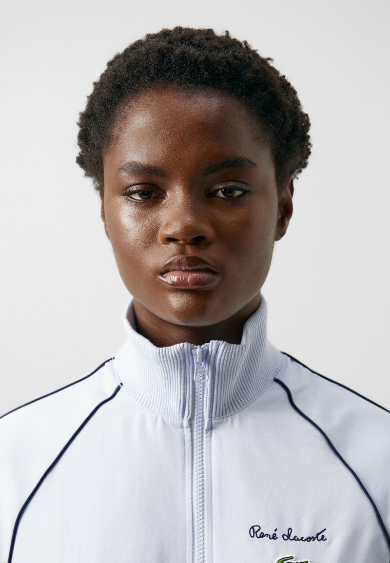 Weiße Zip-Jacke mit hohem Kragen, gerippter Textur und blauen Akzentlinien. Besitzt ein gesticktes Logo auf der linken Brust.