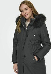 Parka noire avec capuche en fourrure, fermetures à boutons et à zip, taille élastique et poches latérales. Matériau lisse avec une coupe ajustée.