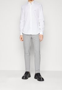 Hombre vestido con una camisa blanca de botones con el logo de CK, pantalones ajustados gris claro y zapatos de cuero negros con cordones, de pie frente a un fondo blanco.