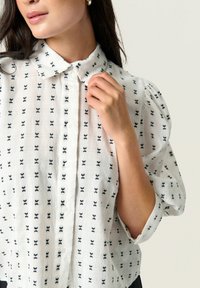 Chemise blanche à boutons avec des motifs de nœuds papillon bleu marine. Dotée d'un col classique et de manches bouffantes, fabriquée en tissu texturé.