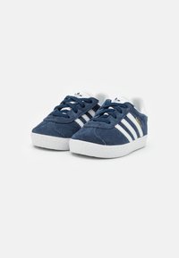 Navy blue suede shoes with white rubber soles, featuring three white stripes and a white tongue with blue laces. Gold logo detail on the side.