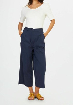 Donna che indossa una maglietta bianca a maniche corte, pantaloni larghi a vita alta di colore blu navy tagliati alla caviglia e sandali con cinturino di colore giallo senape.