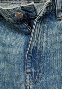 Jean en denim avec une fermeture éclair ouverte et un bouton argenté à la taille, montrant des coutures détaillées et la texture du tissu.