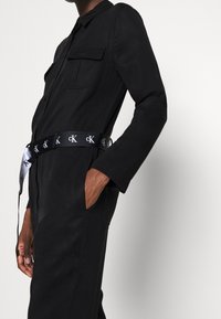 Combinaison noire à manches longues, avec deux poches poitrine, cintrée par une ceinture logo noire et argent. Texture lisse, design moderne.