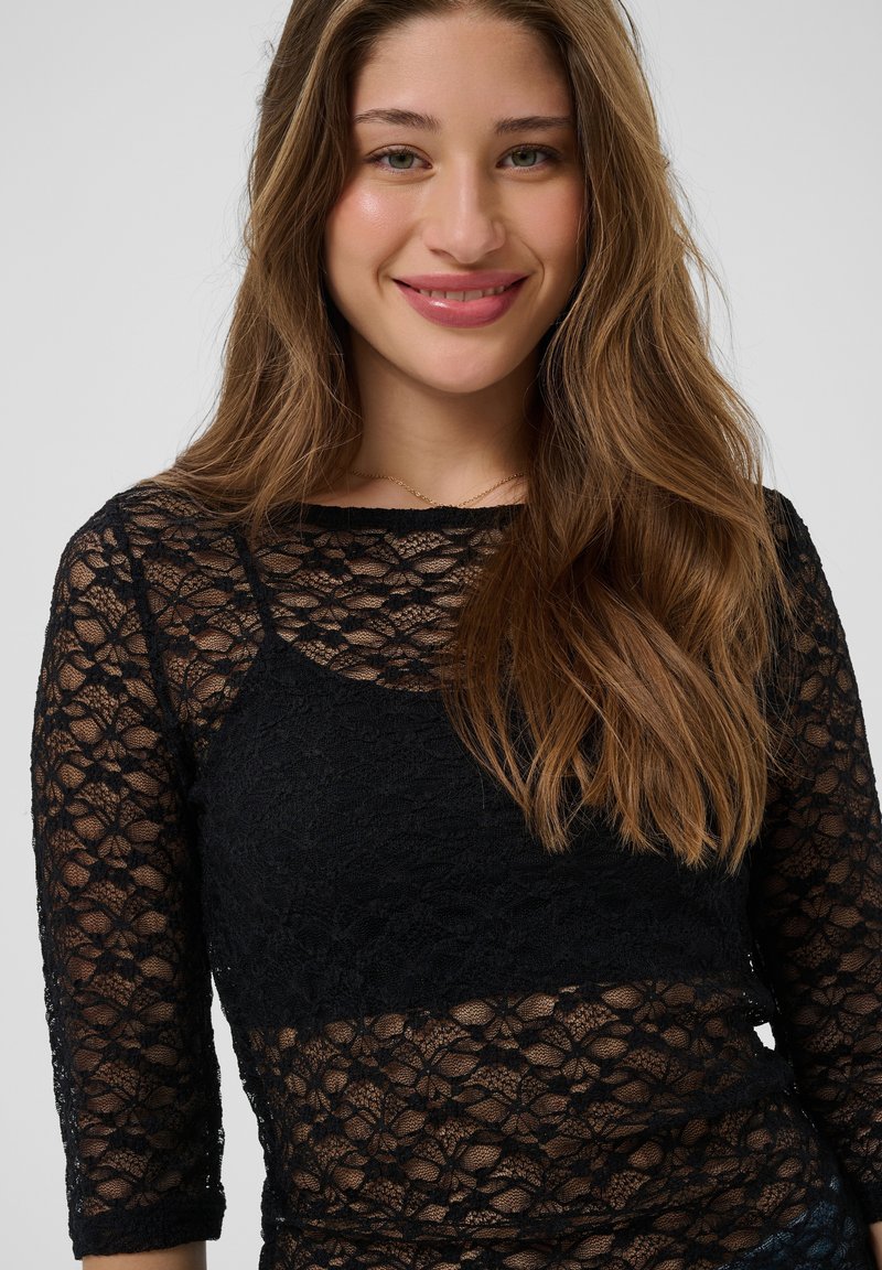 Femme souriante aux longs cheveux bruns portant un haut en dentelle noire à motifs floraux avec des manches trois quarts sur un fond uni.