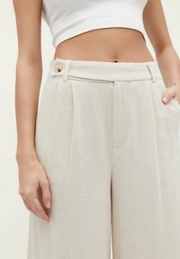 Pantalones de lino beige con un acabado texturizado, que cuentan con una cintura alta, bolsillos laterales y un botón decorativo en la parte frontal de la cinturilla.