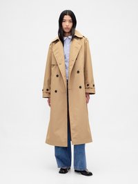 Beige trenchcoat med dobbeltknappet design, stor krage og knappeakenter. Stylet over en stripete skjorte og vide jeans.