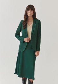 Zelený blazer s jedním knoflíkem, nošený přes béžový pletený top. Oblečení doplňuje odpovídající zelená sukně s volným lemem.