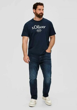 Man with beard wearing navy s.Oliver t-shirt, dark blue jeans, and white sneakers, standing with one hand in pocket against gray background.