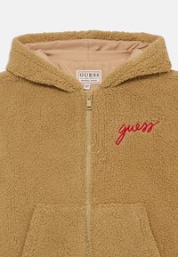 Tanan bunda od flisa s patentnim zatvaračem, koja ima podstavljenu kapuljaču, dva prednja džepa i crveni izvezeni "guess" logo s lijeve strane.