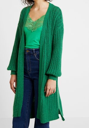 Femme portant un cardigan côtelé vert sur un haut vert orné de dentelle et un jean bleu foncé taille haute, debout devant un fond uni.