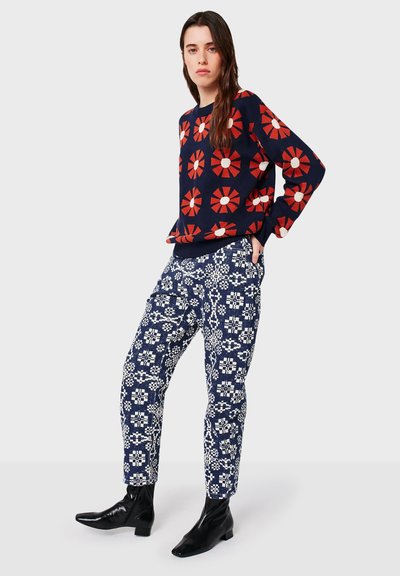 Vrouw in een marineblauwe trui met grote rode en witte bloemen, gecombineerd met marineblauwe broek met een wit geometrisch patroon, en zwarte enkellaarsjes.