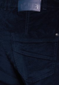 Marinblå kordflodbyxor med en texturerad yta, bakficka och ett lädermärke på midjan. Sydda detaljer är synliga.