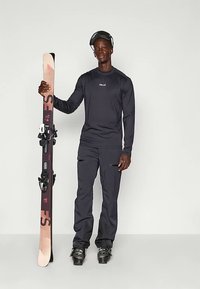 Crna majica s dugim rukavima, crne skijaške hlače i skijaške čizme. Drži skiju s dizajnom u nijansama, jasno vidljiva pričvrsna oprema. Sivo ozadje.