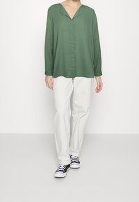 Blusa verde a maniche lunghe con scollo a V e chiusura con bottoni, abbinata a pantaloni bianchi e sneaker nere. Tessuto liscio e vestibilità comoda.