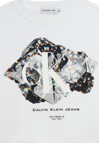 Felpa bianca con un motivo geometrico nero e di cristallo, con "CK" in lettere bianche in grassetto e "Calvin Klein Jeans" stampato sotto.