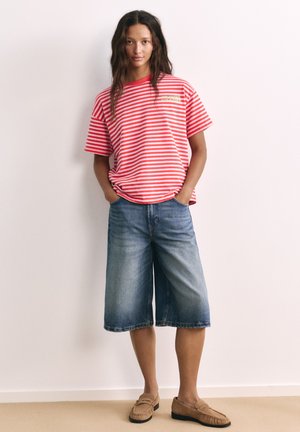 Femme aux longs cheveux portant un t-shirt rayé rouge et blanc, un short en jean arrivant aux genoux et des mocassins couleur crème, debout contre un mur blanc.