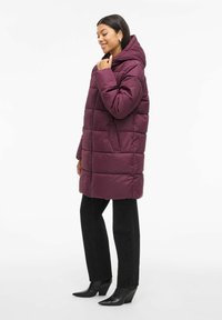 Pufasta jakna u tamno burgundskoj boji s visokim ovratnikom i zatvaranjem na patentni zatvarač. Ima quilted dizajn i dva bočna džepa. Nosena je s crnim hlačama.