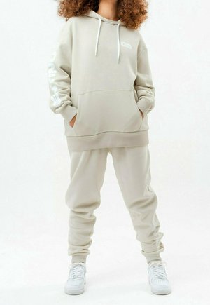 Person med krøllet hår iført beige oversized hættetrøje og joggingbukser med hvide sneakers, der står med hænderne i lommerne mod en hvid baggrund.