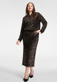 Schwarzes, mit Pailletten besetztes zweiteiliges Outfit mit einem Reißverschluss-Jacke mit Bündchen und einem passenden langen Rock. Hochhackige Schuhe vervollständigen das Outfit.