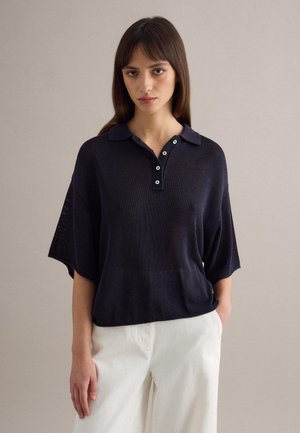 Mujer con cabello liso y castaño, vestida con una camiseta polo de manga corta suelta color azul marino oscuro y pantalones blancos, sobre un fondo claro y simple.