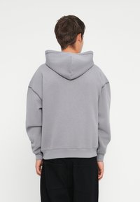 Pelēks kokvilnas kapuce, kas raksturojas ar lielu kapuci, nolaižamām plecu šuvēm un ribotām aprocēm. Mugura ir vienkārša, bez redzamām logo vai rakstiem.