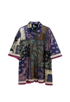 Joven de pie con las manos en los bolsillos, vistiendo una camisa de manga corta de patchwork multicolor, pantalones rosas a cuadros y zapatos beige texturizados.