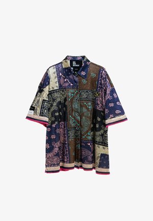 Joven de pie con las manos en los bolsillos, vistiendo una camisa de manga corta de patchwork multicolor, pantalones rosas a cuadros y zapatos beige texturizados.
