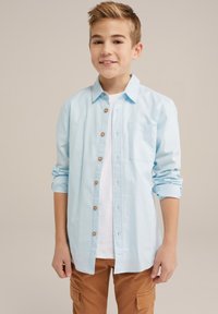 Svetle modrá košile na knoflíky s kapsou na hrudi, ohrnutými rukávy a hnědými knoflíky. Nositá přes bílou košili a béžové cargo kalhoty.