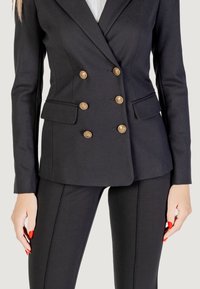 Donna che indossa un blazer nero aderente con bottoni dorati e pantaloni neri abbinati, le mani con smalto rosso riposano ai lati.