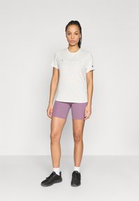 Ανοιχτό γκρι μπλουζάκι με το λογότυπο "Champion", σε συνδυασμό με μωβ ποδηλατικά σορτς και μαύρα αθλητικά παπούτσια, σε μονόχρωμο φόντο.