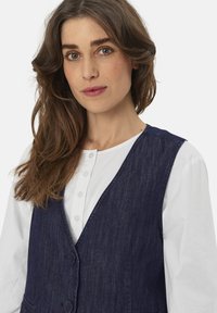 Dunkelblaue Denim-Weste mit V-Ausschnitt, mit Knopfverschluss und Taschen, über einem weißen Langarmhemd getragen. Glatte Textur.