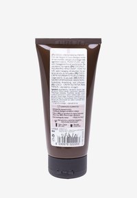 Brun plasttube med handkräm och svart lock, märkt med flera språk och ingredienser. Texturerad yta med glänsande accenter.