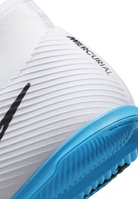 Valkoinen urheilukenkä, jossa on teksturoitu yläosa, musta logo ja sininen jälkiä jättämätön pohja, suunniteltu ketteryyteen sisätiloissa.